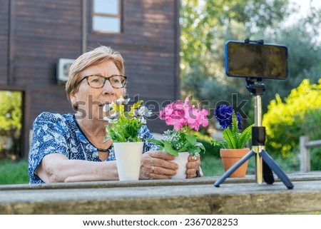 Senior woman recording a podcast and vlogging with smartphone sitting on a park bench, with flowers on the table.