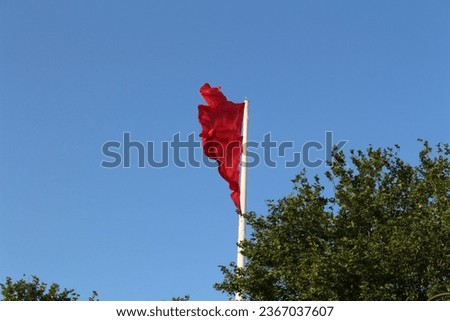 The photo is taken from below, featuring a white pole with a waving red flag as the focal point. Behind, there's a blue sky, and to the right of the photo, a tree with green leaves can be seen.