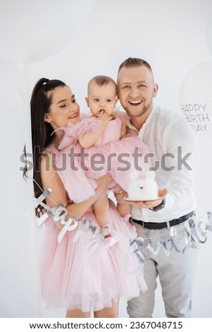 Blurred background of caucasian family of mother, father and little daughter posing in embrace among festive decoration in studio. Positive man holding cute birthday cake in form of teddy bear.