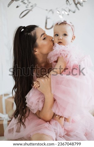 Happy caucasian mom kissing cheeks of adorable daughter among birthday decor and presents in white studio. Positive woman in stylish evening dress celebrating anniversary of her lovely little girl.