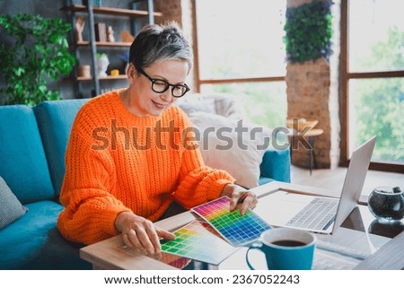 Photo of smiling cheerful mature lady dressed orange sweater choosing color design modern gadget indoors house room