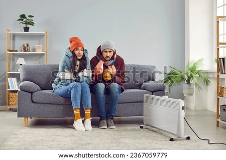 Freezing people in winter jackets sitting on sofa at home. Sad warmly closed husband abd wife feeling cold and trying to warm up. Heating problems, power crisis concept Royalty-Free Stock Photo #2367059779