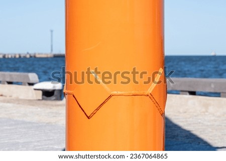 A huge metal bright orange tube on the seashore. In the background, a light pavement, a wooden fence and the sea. Copy space. Background for quotes. High quality photo