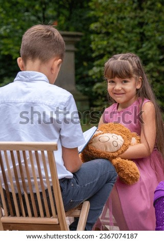 The boy sits with his back on a chair and reads a book. The girl looks in with interest and wants to look at the pictures. Girl 6 years old, holding a teddy bear, with long dark hair 