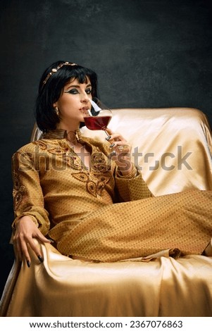 Portrait of noble, beauiful yougn woman in imag of queen, Cleopatra, in goden dress sitting and drinking red wine against dark background. Concept of antique culture, history, comparison of eras, art Royalty-Free Stock Photo #2367076863