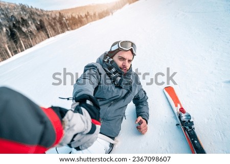 Helping out - giving a hand to a skier that fell down. Man fell to the ground reach out to his friend and trying to get up from the snow. Ski accident on ski slope in the mountains.