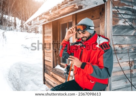 Young smiling male skier using smart phone in front of log cabin. Fashionable man in full ski equipment checking his mobile phone on vacation.