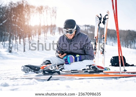Smiling male skier sitting on snow taking a break while using phone and texting. Happy athletic man scrolling his cellphone and feeling satisfied. Royalty-Free Stock Photo #2367098635
