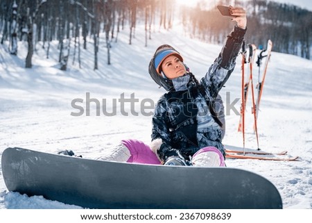 Beautiful female snowboarder sitting on snow and taking a break while taking a selfie. Young athletic woman enjoying winter holidays using mobile phone to make a photo in snow.