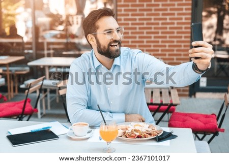 Handsome male person sitting in the restaurant and having a lunch. Smiling man is holding a phone and taking a selfie outdoors.