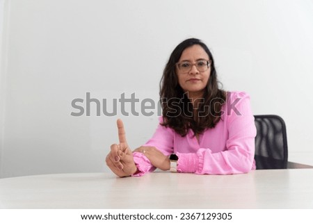 Portrait of a psychopedagogue, wearing pink clothes, in her office, sitting, making a sign with her hand. Helps with school performance. Take care of your mental health.