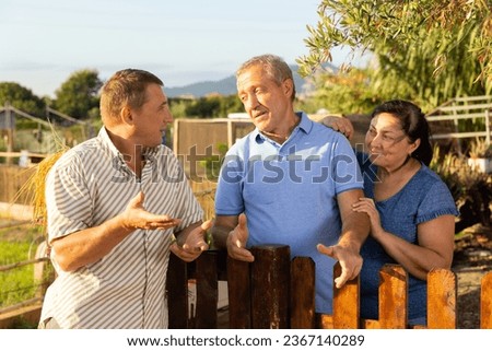 Man standing at fence and talking with mature couple neighbours at backyard garden. Royalty-Free Stock Photo #2367140289
