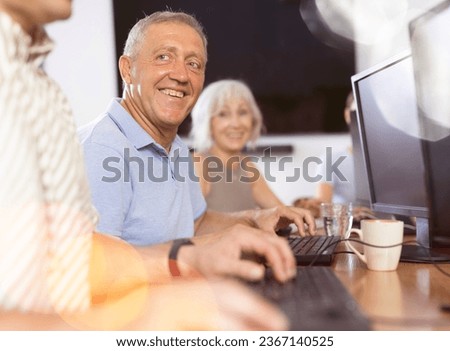 Smiling motivated older man engrossed in exploring digital realm at internet cafe, using computer, demonstrating curiosity of modern seniors towards technology..