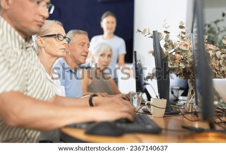 Interested senior woman in glasses attending computer class, learning basics of digital technology under guidance of teacher standing in blurred background
