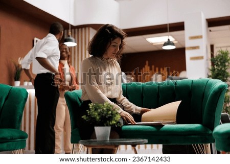 Female manager checking bureaucracy to review booking forms, sitting on couch in lounge area. Young hotel concierge working on registration details, providing luxury resort service. Royalty-Free Stock Photo #2367142433