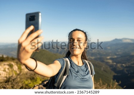 A young, happy woman smiles as she takes a selfie with her smartphone from the top of a mountain