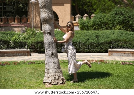 Young and beautiful blonde and latin woman leans on the trunk of a palm tree in the park doing different poses while having her picture taken. The woman is happy and having fun on a sunny day