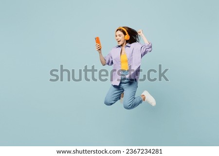 Full body side view young happy woman she wears purple shirt yellow t-shirt casual clothes jump high use mobile cell phone listen to music in headphones isolated on plain pastel light blue background