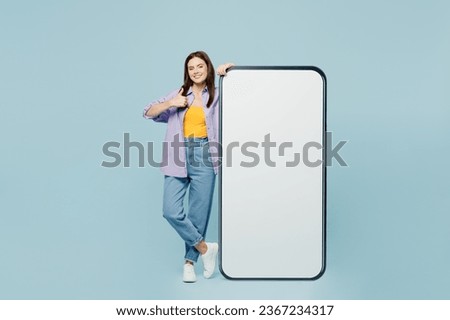 Full body young fun woman wears purple shirt yellow t-shirt casual clothes big huge blank screen mobile cell phone smartphone with area show thumb up gesture isolated on plain light blue background