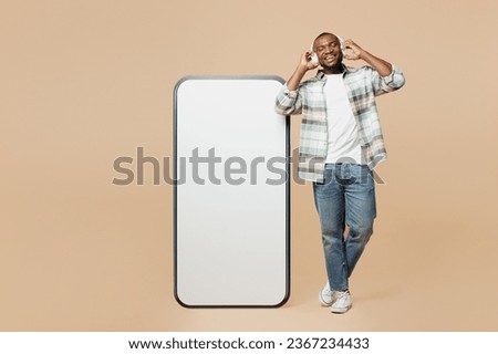 Full body young man he wear light shirt casual clothes big huge blank screen mobile cell phone with workspace copy space mockup area listen to music in headphones isolated on plain beige background