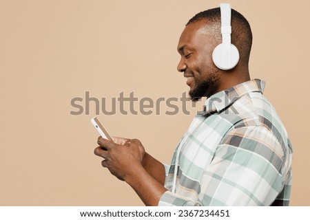 Side view young happy man of African American ethnicity wear light shirt casual clothes listen to music in headphones use mobile cell phone isolated on plain pastel beige background. Lifestyle concept