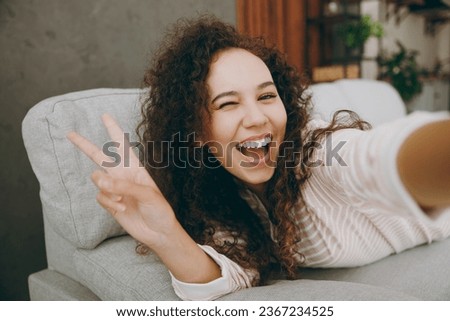 Close up young woman wear casual clothes lay down on grey sofa couch doing selfie shot pov mobile cell phone show v-sign stay at home hotel flat rest relax spend free spare time in living room indoor