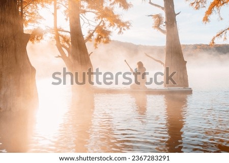 Woman relaxing on staand up paddle board at quiet lake with morning fog and fall Taxodium distichum trees Royalty-Free Stock Photo #2367283291