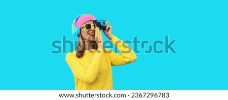 Stylish modern young woman photographer taking a picture with film camera and listens to music in headphones wearing colorful pink hat, yellow sweater on blue studio background