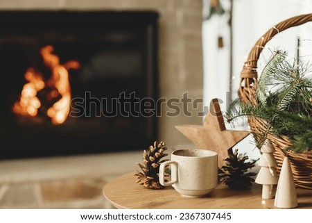Winter cozy living room in farmhouse. Stylish cup of warm tea, basket with fir branches, wooden trees and star, pine cones against burning fireplace. Modern christmas rustic eco friendly decor