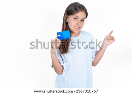 Smiling young beautiful kid girl wearing wearing grey T-shirt showing debit card pointing finger empty space
