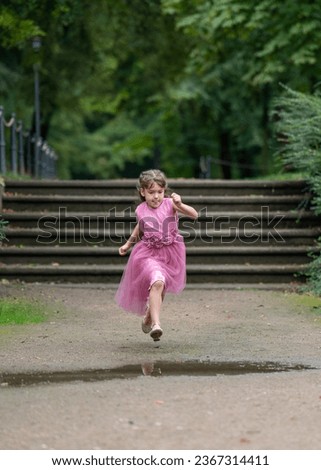 A little girl in a beautiful pink dress with long hair runs quickly and jumps over a puddle. I concentrated and bit my tongue. Outdoor games. Blurred background. 
