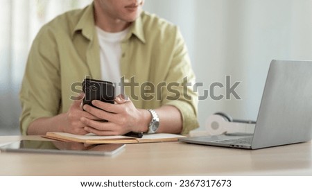 Cropped image of a handsome Asian man in casual clothes is holding his smartphone while looking at something on his laptop at his desk.