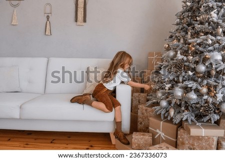 Little blonde girl in stylish, fashion mustard, corduroy pants, white shirt decorates rustic Christmas tree. Toddler sits on sofa, background of gray wall with macrame. Trendy interior. Copy space