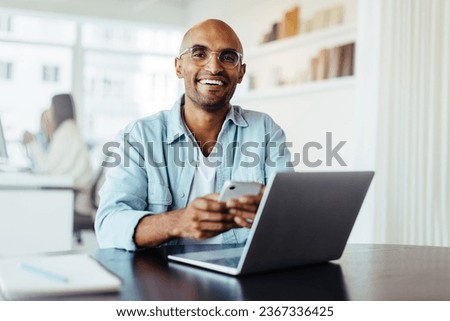 Business man smiling at the camera while holding a mobile phone. Young male designer sitting in an office with a laptop.