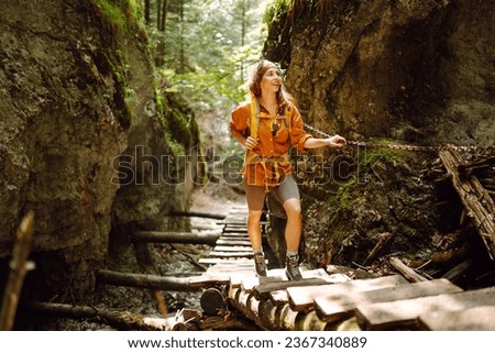 Happy woman in hiking clothes with a yellow backpack walks along a wooden hiking path in the mountains. Hiking, active lifestyle. Outdoor adventure concept.