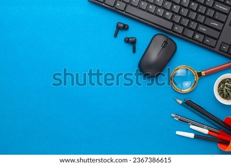 Work desk with black keyboard, mouse, earbuds, magnifying lens, pen, pencil and plant on a Blue Background. Top-down view.