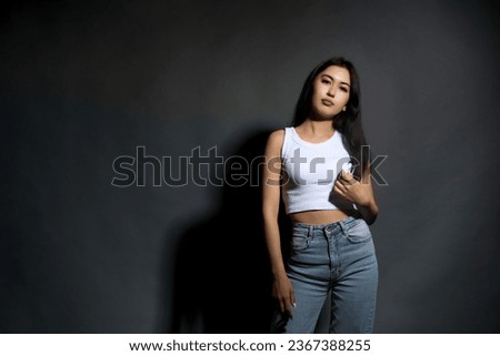 Thoughtful, calm, slender young woman in jeans and a white short T-shirt poses dreamily against a gray background. Stylish long-haired girl in modern youth clothes in the studio at a photo shoot