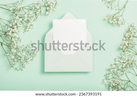 Delicate white gypsophila flowers, a light green envelope and a blank card on a green background. Flat composition. Postcard, invitation.