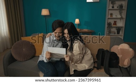 Photo of a teenager girl, young woman, daughter sitting with her mother. They are reading the paper, letter, document, excited with what they're seeing.