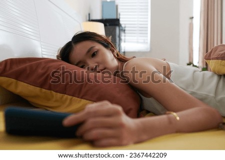 Tired depressed young woman lying in bed and watching show on smartphone