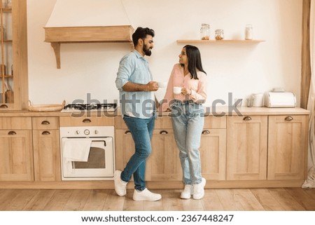 Happy relaxed indian spouses with cups enjoying coffee and free time together in cozy kitchen interior, full length, free space. Couple relationship, love and breakfast
