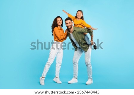 Cheerful european family of three having fun, dad piggybacking daughter over blue background, parents and their child smiling at camera, full length, free space