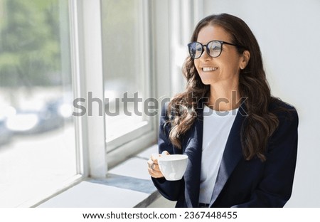 Successful cheerful attractive mature businesswoman in formal outfit wearing eyeglasses standing next to window with mug in her hand, drinking coffee at office, enjoying view, copy space Royalty-Free Stock Photo #2367448455