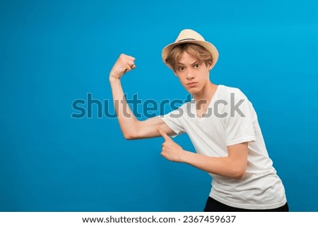 Look at my bicep. Picture of funny smiling teen boy in casual white t-shirt posing isolated at bright color blue background, showing his arm muscle. Childhood, fitness and sports concept