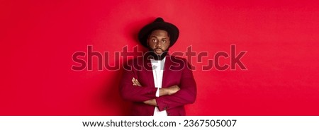 Serious black man standing in party outfit and waiting, cross arms on chest staring at camera, red background.