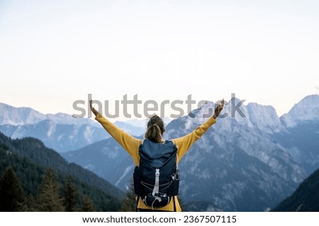 View from behind of a female hiker with a backpack standing with her arms spread wide looking at a beautiful view of high mountains under clear sky. Royalty-Free Stock Photo #2367507115