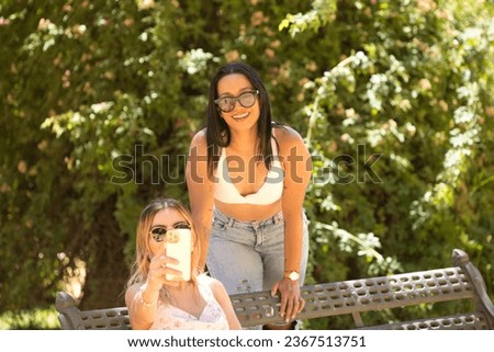 Two young and beautiful latin american women on holiday take selfie very happy. One is sitting and the other one is leaning on the bench. Concept of friendship and travel
