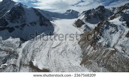 An ancient snow glacier among high mountains. The peaks of the mountains are covered with snow. In places there are steep cliffs and large rocks. Blue sky with white clouds. Top view from a drone