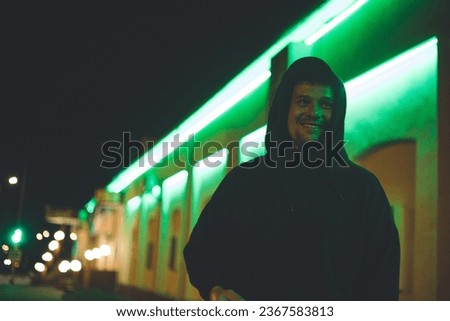 On the street of a city at night, a handsome man laughs. A man in a hood smiles sweetly at night in the neon light of the city. City nightlife concept. Royalty-Free Stock Photo #2367583813