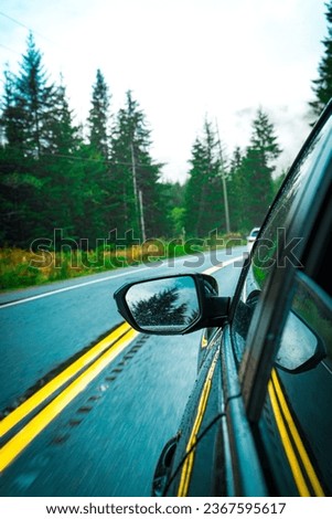 A vertical shot of a car driving on a paved road bend with a forest and the sky in the background.